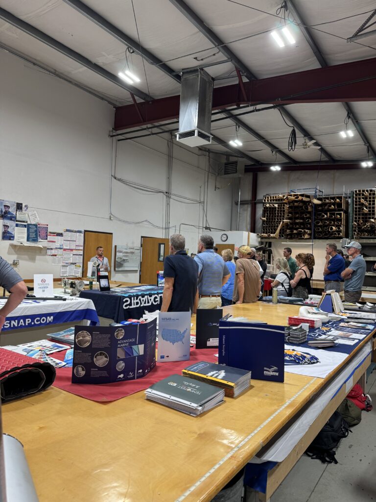 Inside a fabrication shop with a long table displaying brochures, catalogs and promotional products; attendees watch a presenter give a demonstration.