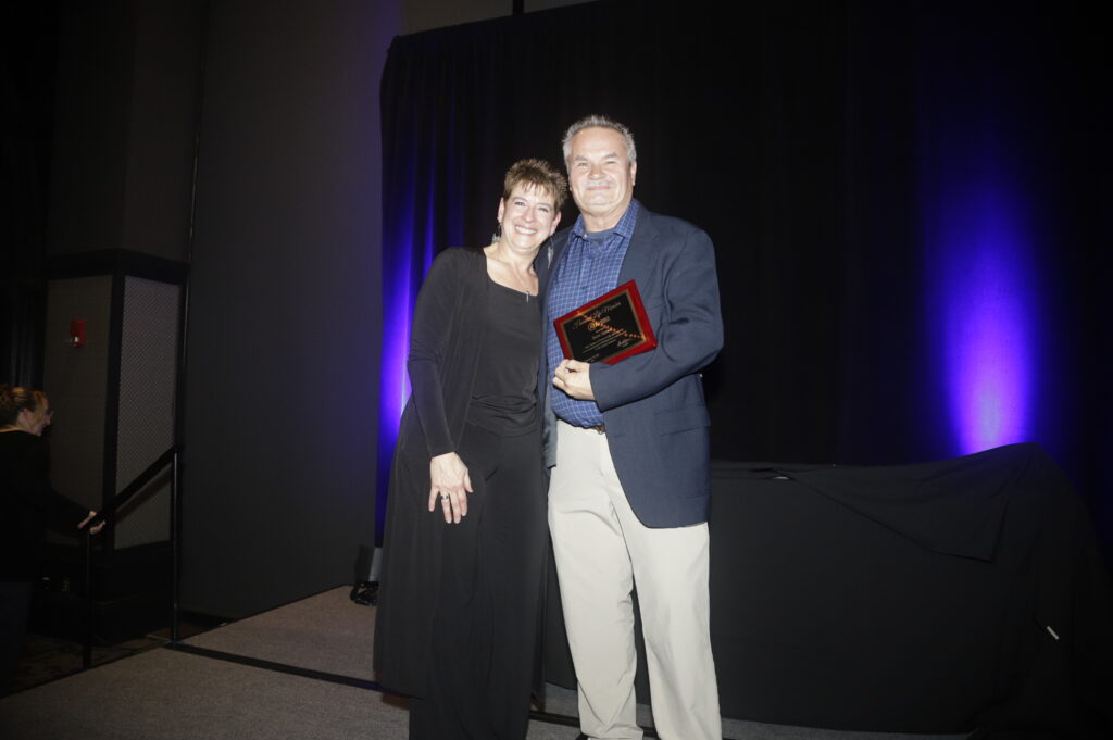 Two individuals pose together on stage, one holding an award, with a dark backdrop illuminated by purple lighting.