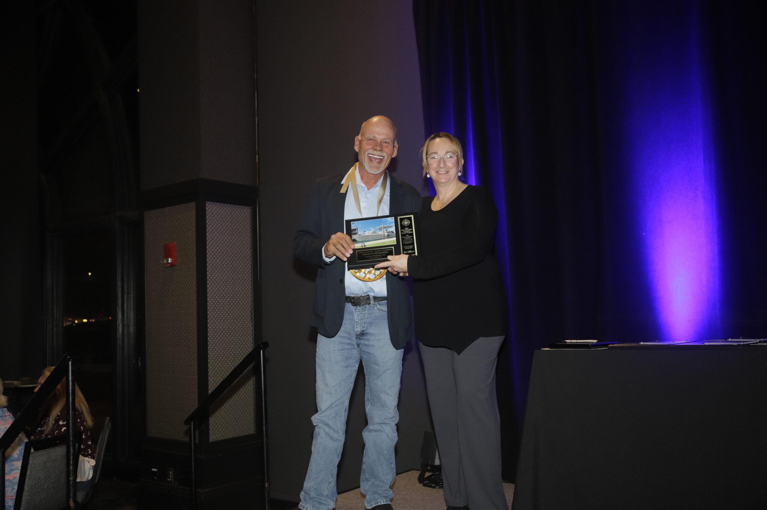 A man and woman smile as they pose with an award plaque, set against a backdrop of dark fabric and a purple-lit area.