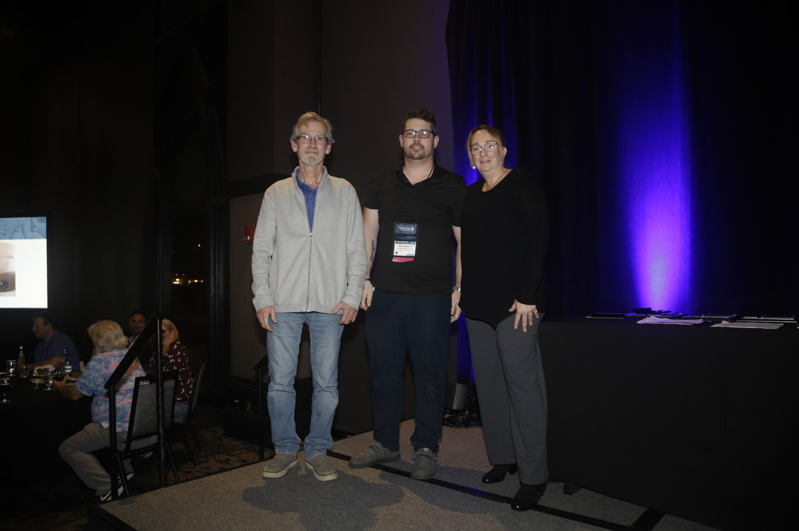 Two award recipients and a presenter stand on a stage in front of a dark backdrop, with purple lighting, against an audience and a table displaying awards.