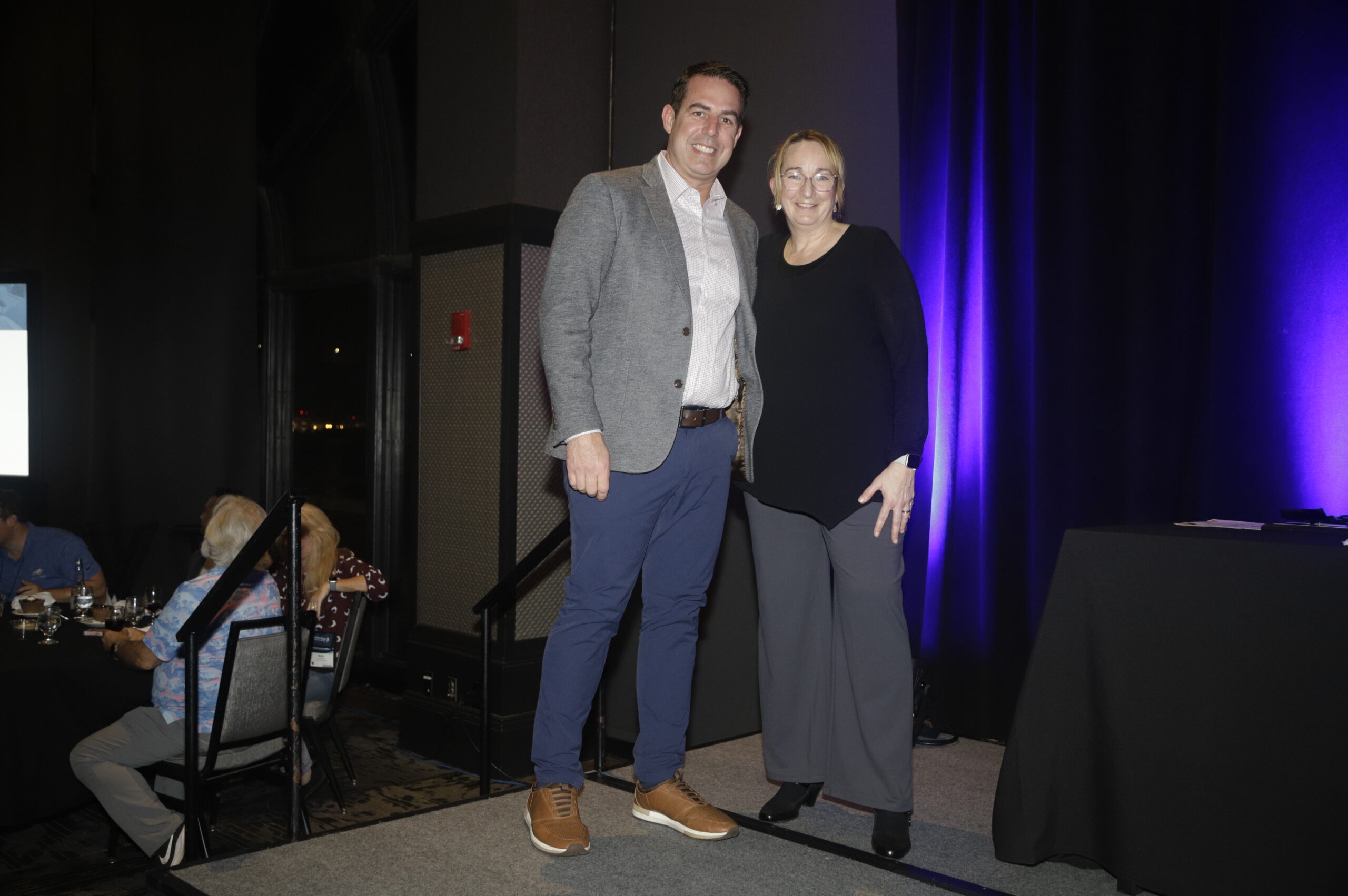 An award winner and a presenter stand on stage, smiling together. The backdrop features purple lighting, with tables set for an event visible behind them.