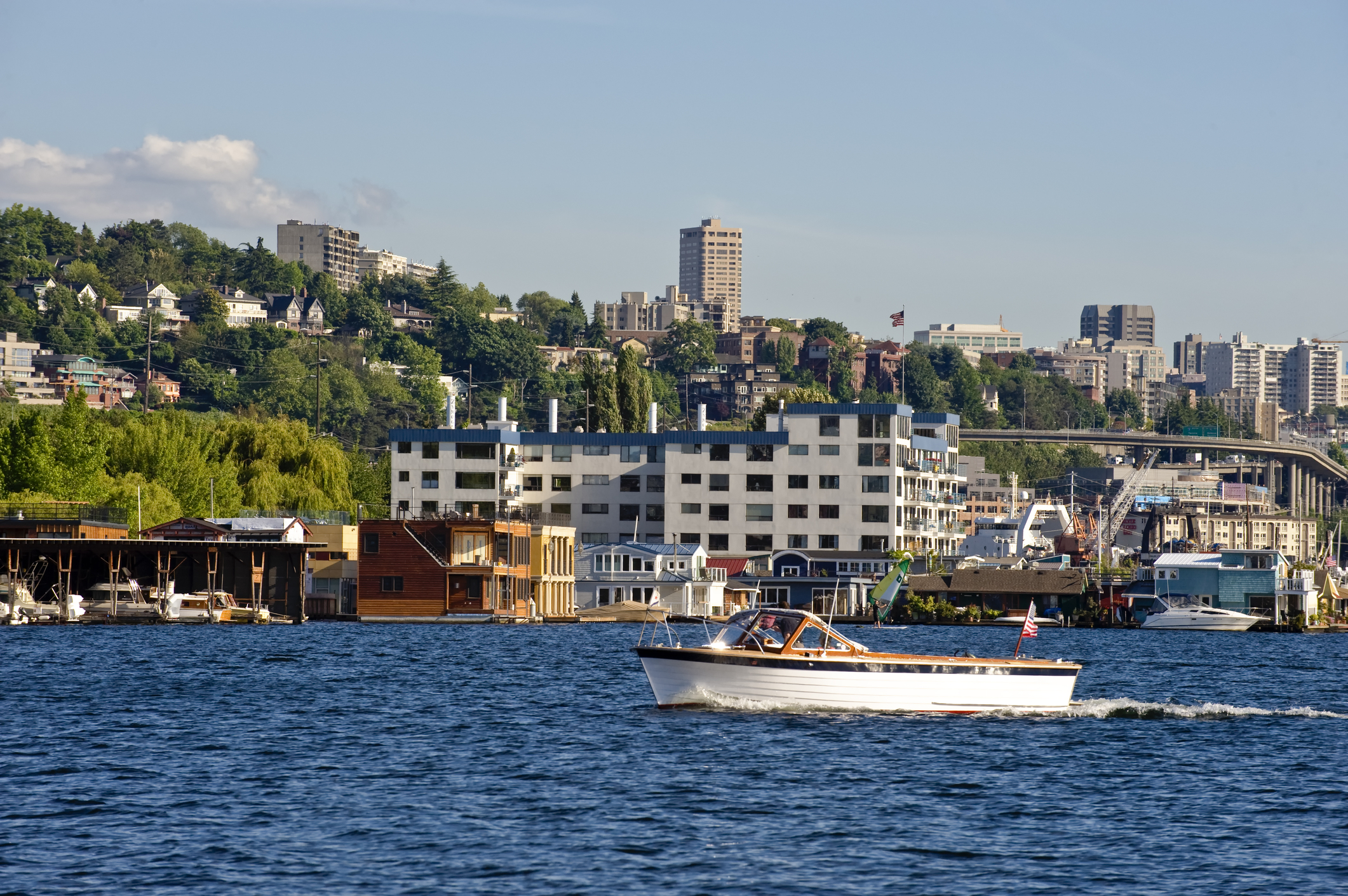 Wood motorboat navigating a harbor, dotted with houseboats and backed by hills and city buildings.