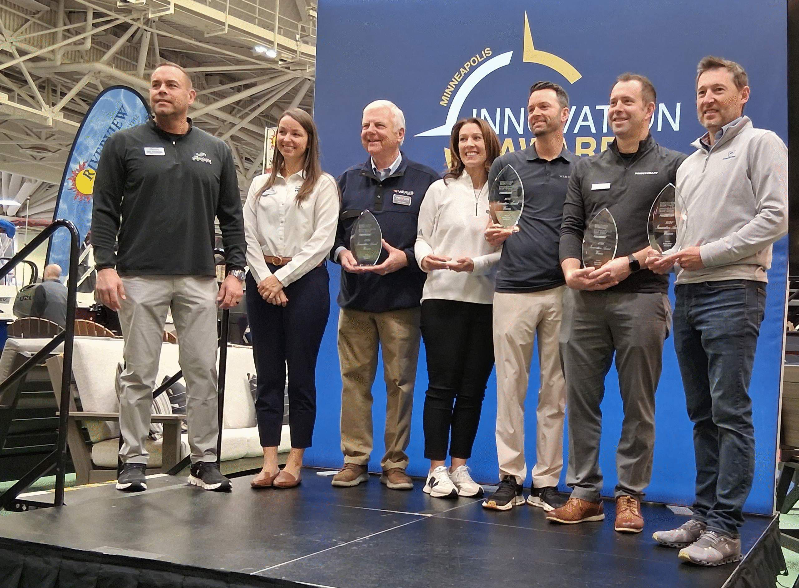 Six award winners stand on a stage, holding trophies, against a blue backdrop with "Minneapolis Innovation Awards" text.