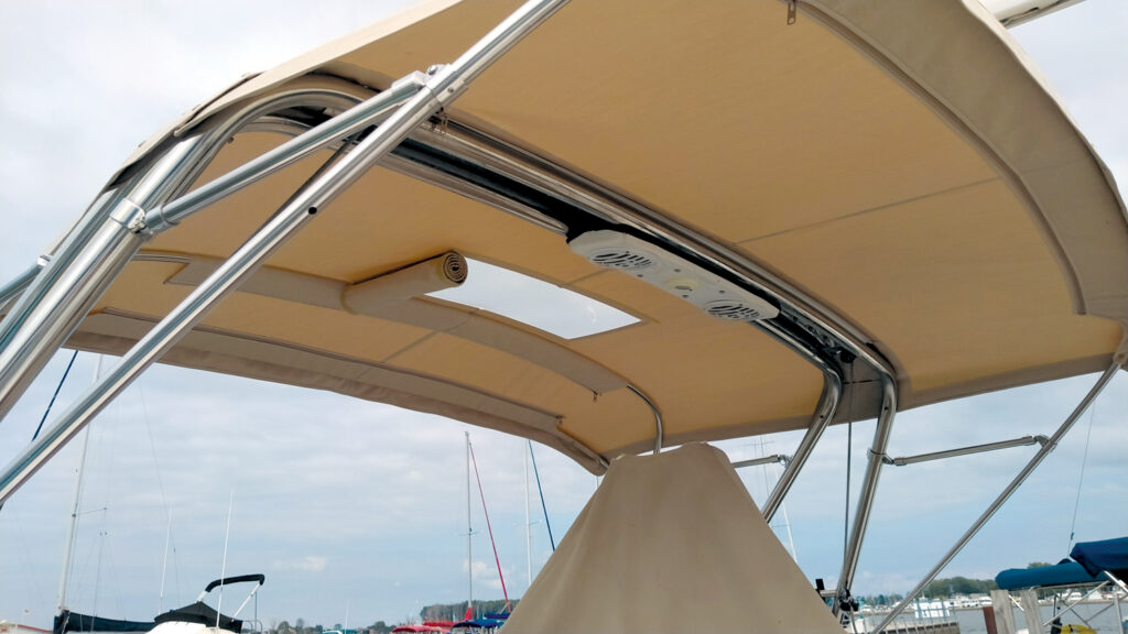 View of a boat's tan canopy and support structure, featuring metallic frames and mounted speakers, with sailboats visible in the background.