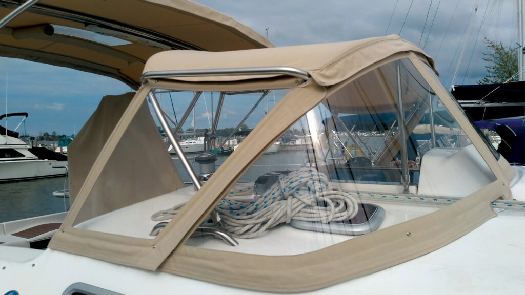 Transparent boat canopy with beige fabric and metal frame, showing arranged ropes on a white deck, marina in the background.