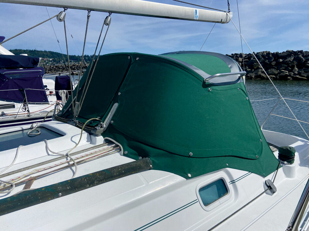 Green fabric cover on a sailboat, with ropes and hardware against a backdrop of water and rocky shore.