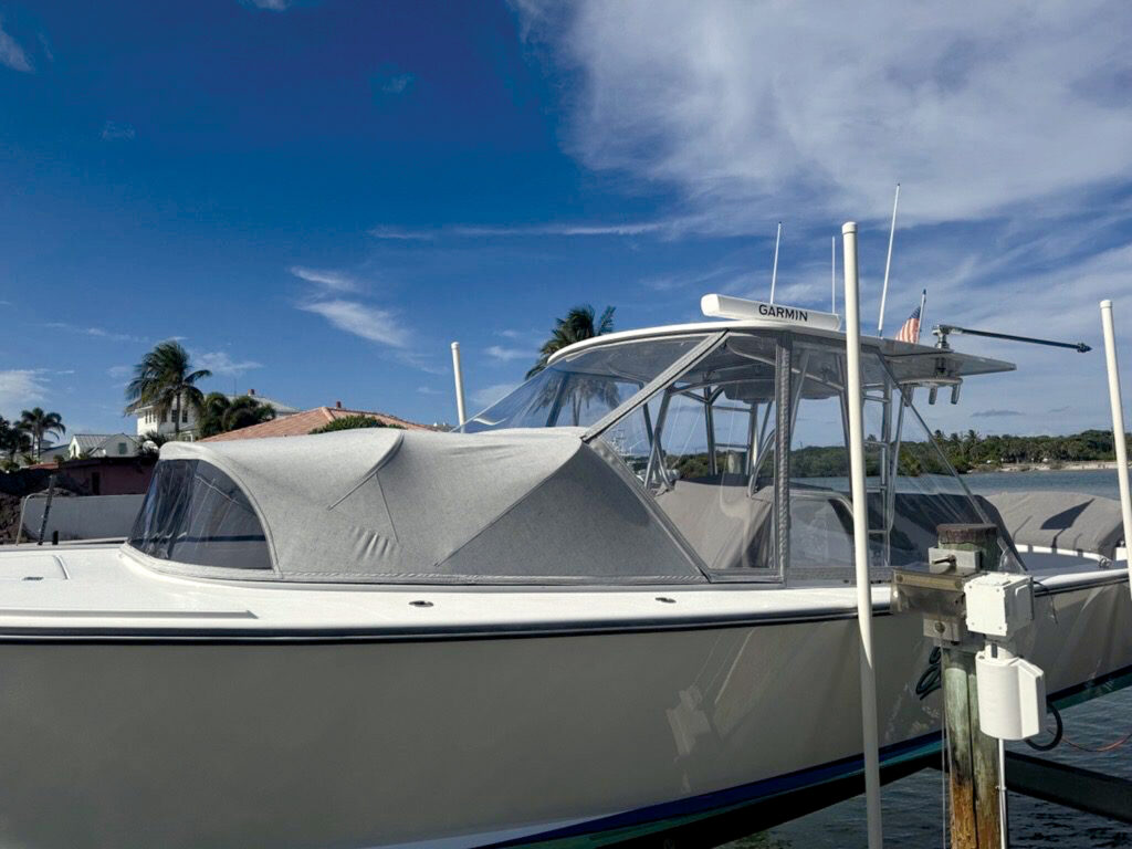 A modern powerboat with a gray cover, docked under a clear blue sky, surrounded by palm trees and residential buildings.