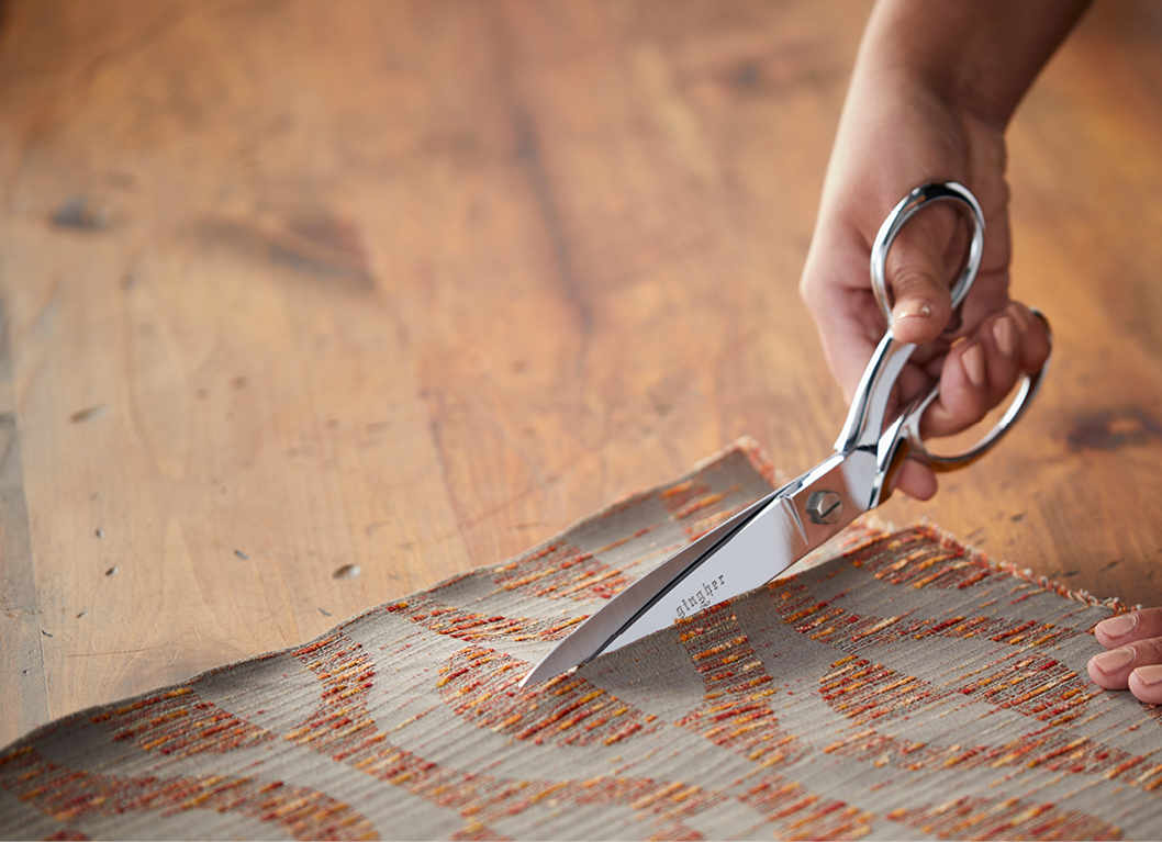 Hand holding a pair of shiny scissors cutting through patterned fabric on a wooden surface.
