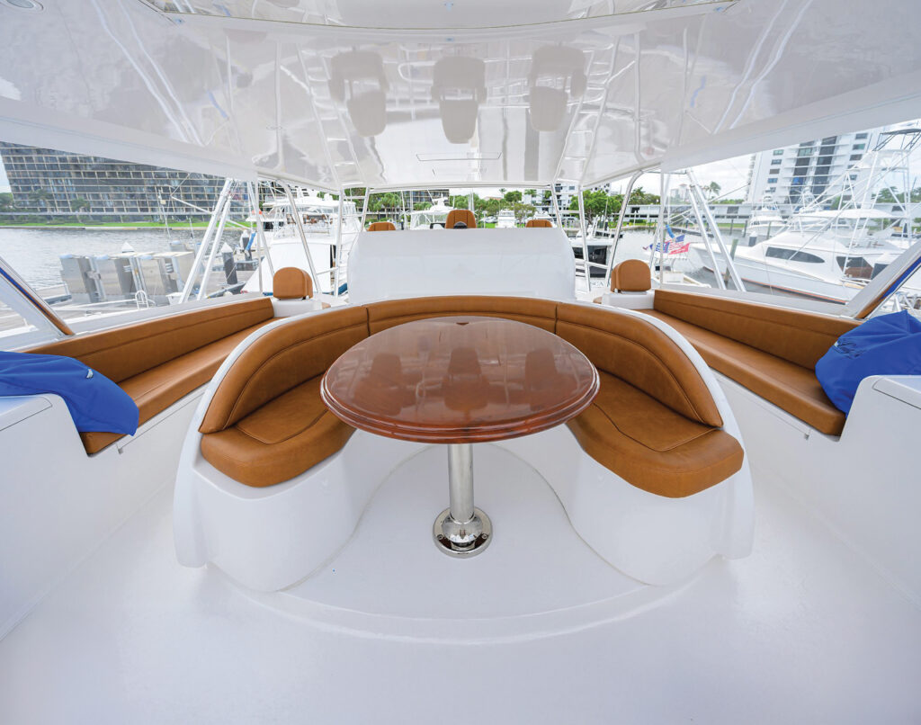 Flybridge seating with curved brown cushions surrounding a polished table, set against a backdrop of boats and marina at sunny waters.