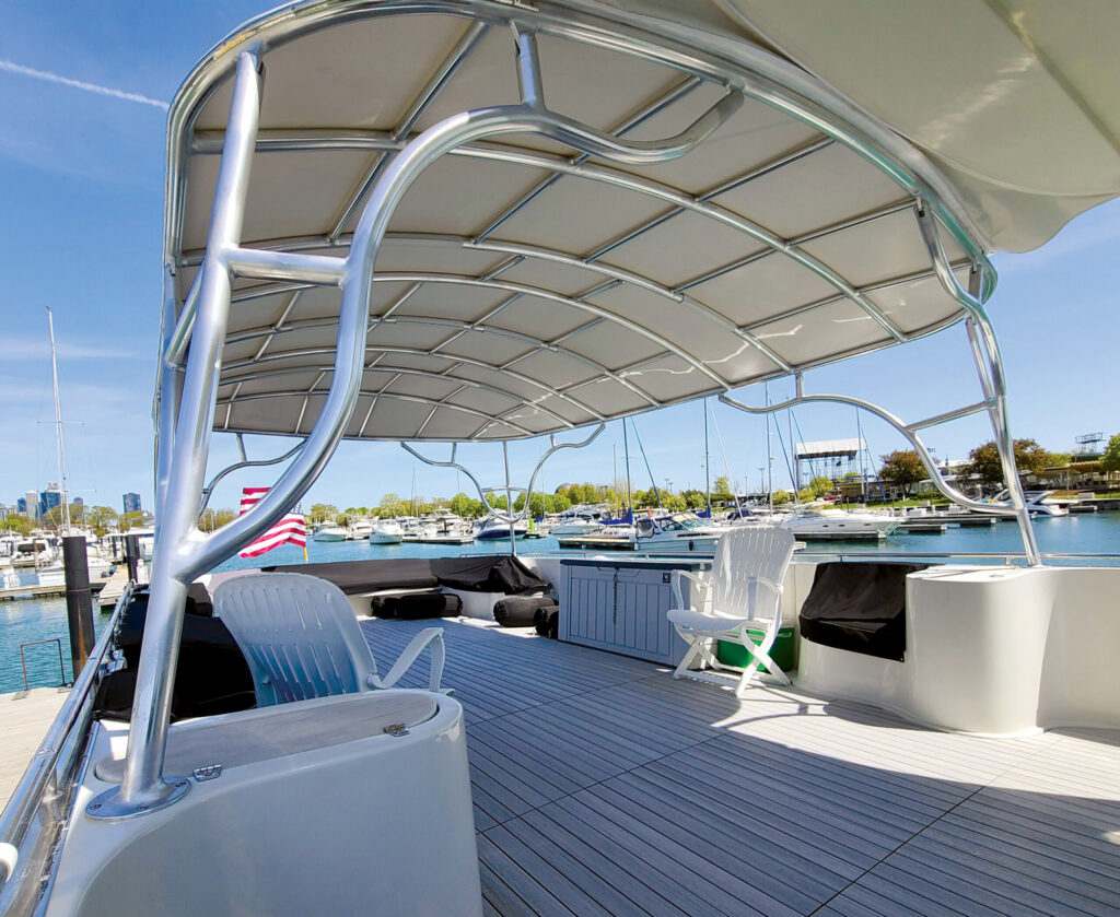 Beneath a curved aluminum frame, a spacious boat deck features white seating, a grey cabin and a scenic view of a marina and city skyline.
