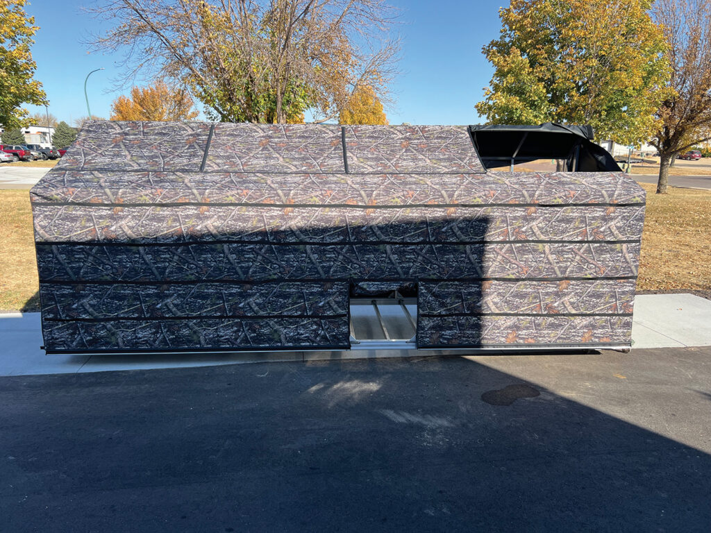 Camo-patterned hunting blind on a paved surface, with autumn trees in the background and a clear blue sky above.
