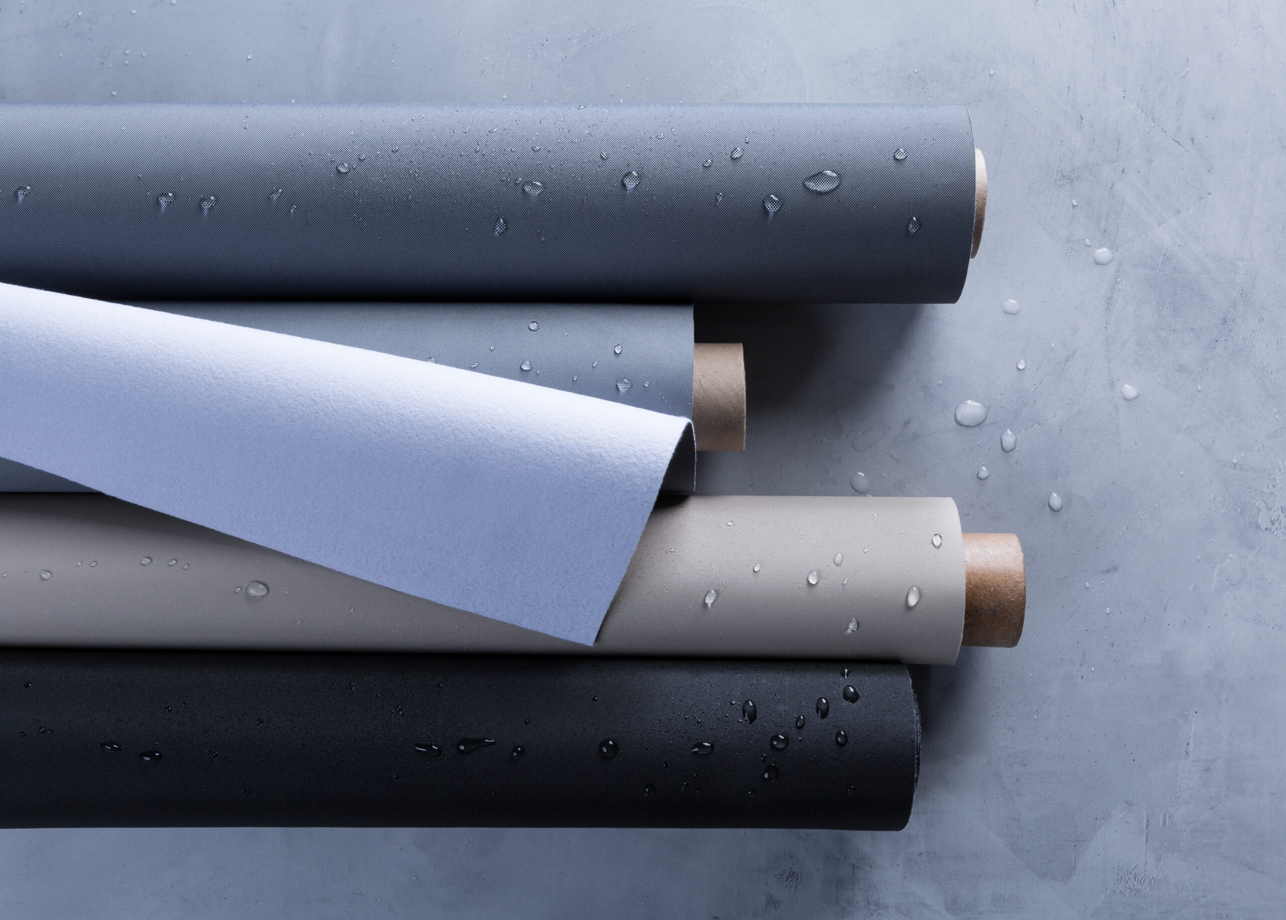 Rolls of textured fabric in shades of gray, beige and white, with water droplets, arranged on a smooth gray surface.