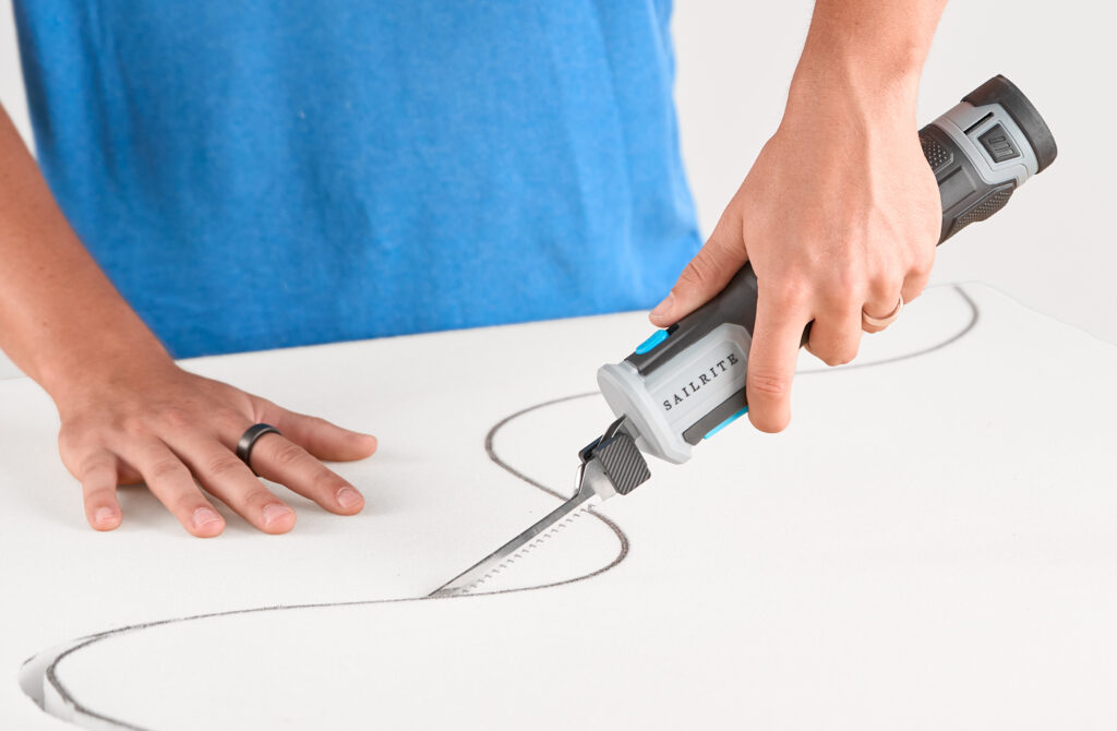 A hand holds a cordless foam saw, cutting a curved line in white foam on a table, with a blue shirt visible in the background.