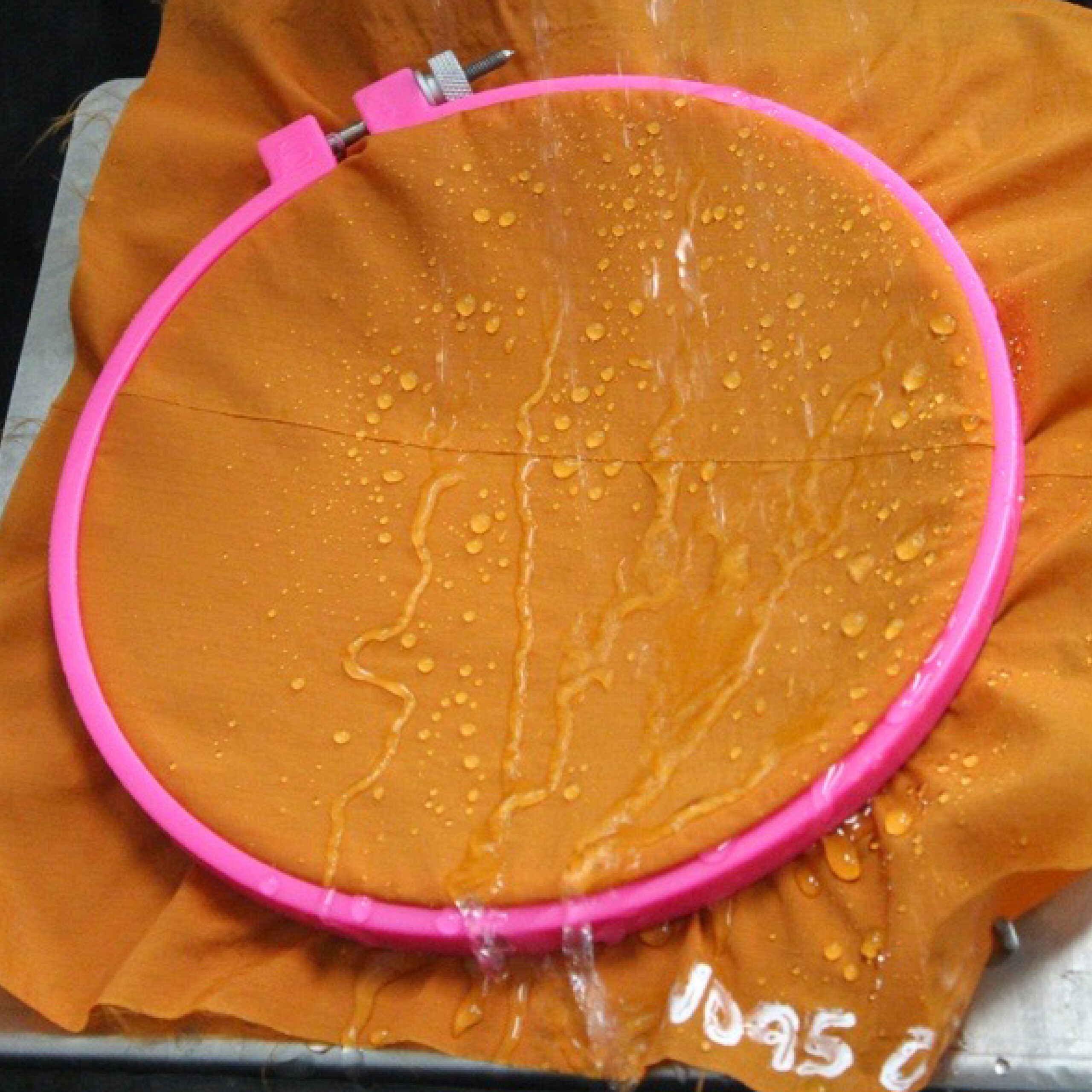 Bright orange fabric with water droplets displayed inside a pink embroidery hoop, highlighting water-resistant qualities in marine materials.