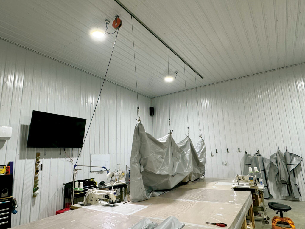 Brightly lit workshop featuring sewing machines, a large table covered with fabric and suspended grey tarps against metal walls.