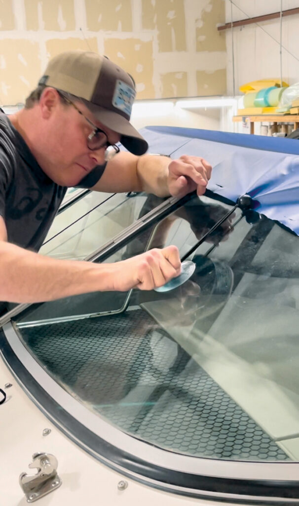 A man in a cap uses a suction tool to install a windshield in a boat, surrounded by a workshop with neutral colors.