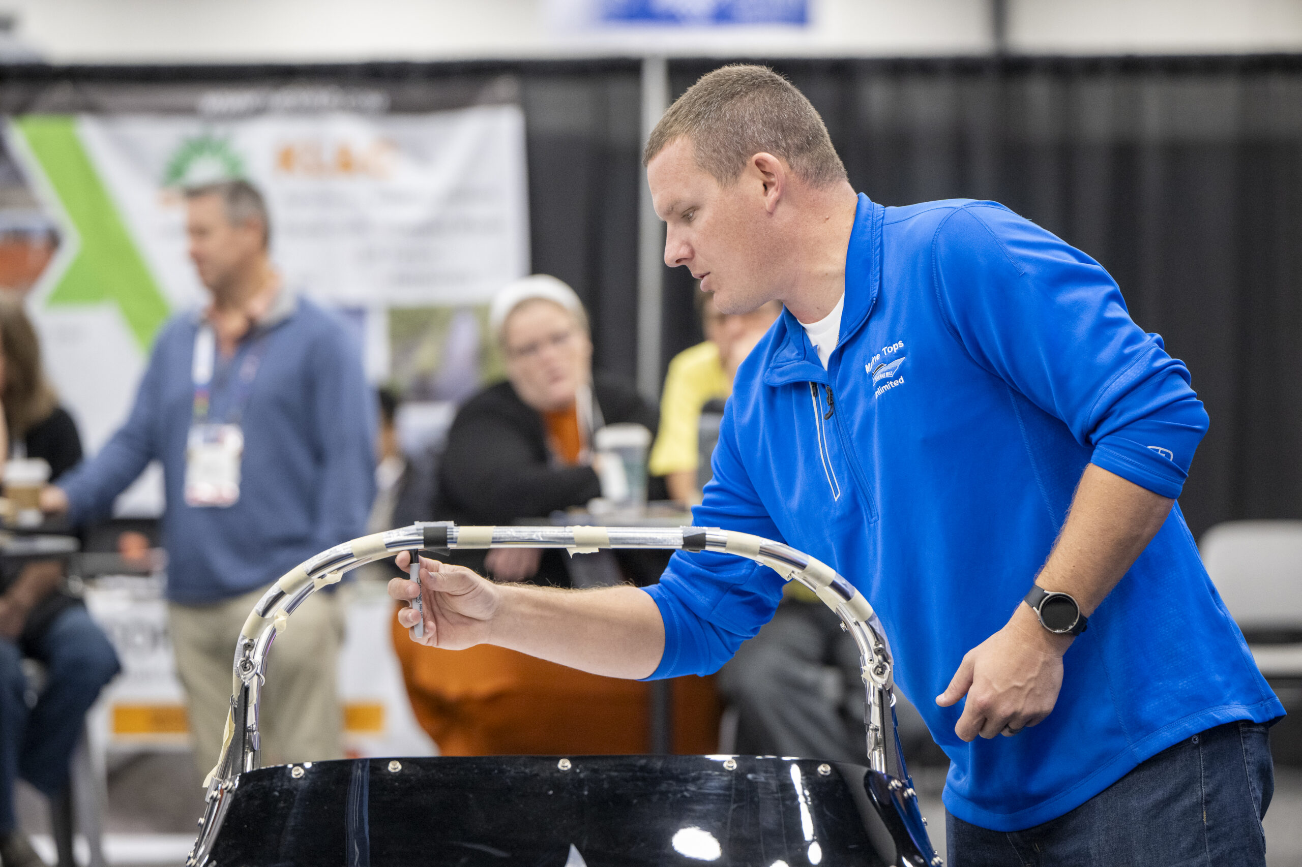 Man in a blue shirt demonstrating marine fabrication on a sleek black surface, with blurred background of attendees and displays.