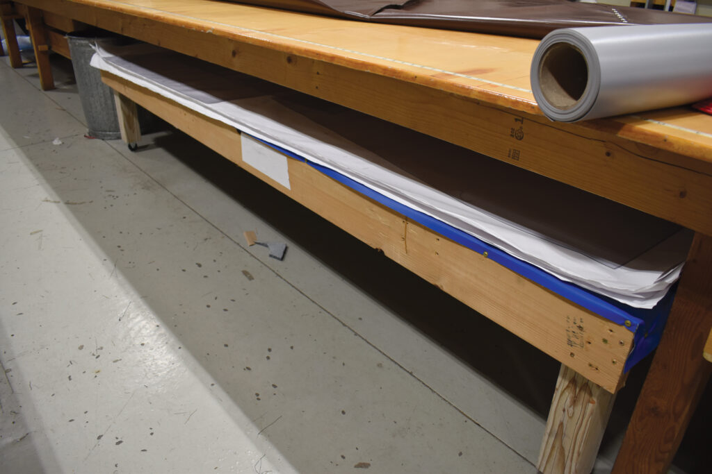 Wooden workbench with a rolled vinyl material atop, and storage beneath holding various sheets, set on a concrete floor.