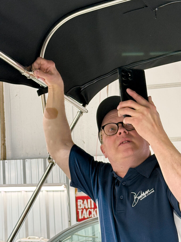 A man in a navy shirt inspects a boat canopy, holding his phone while adjusting a metal frame, illuminated in a workshop setting.