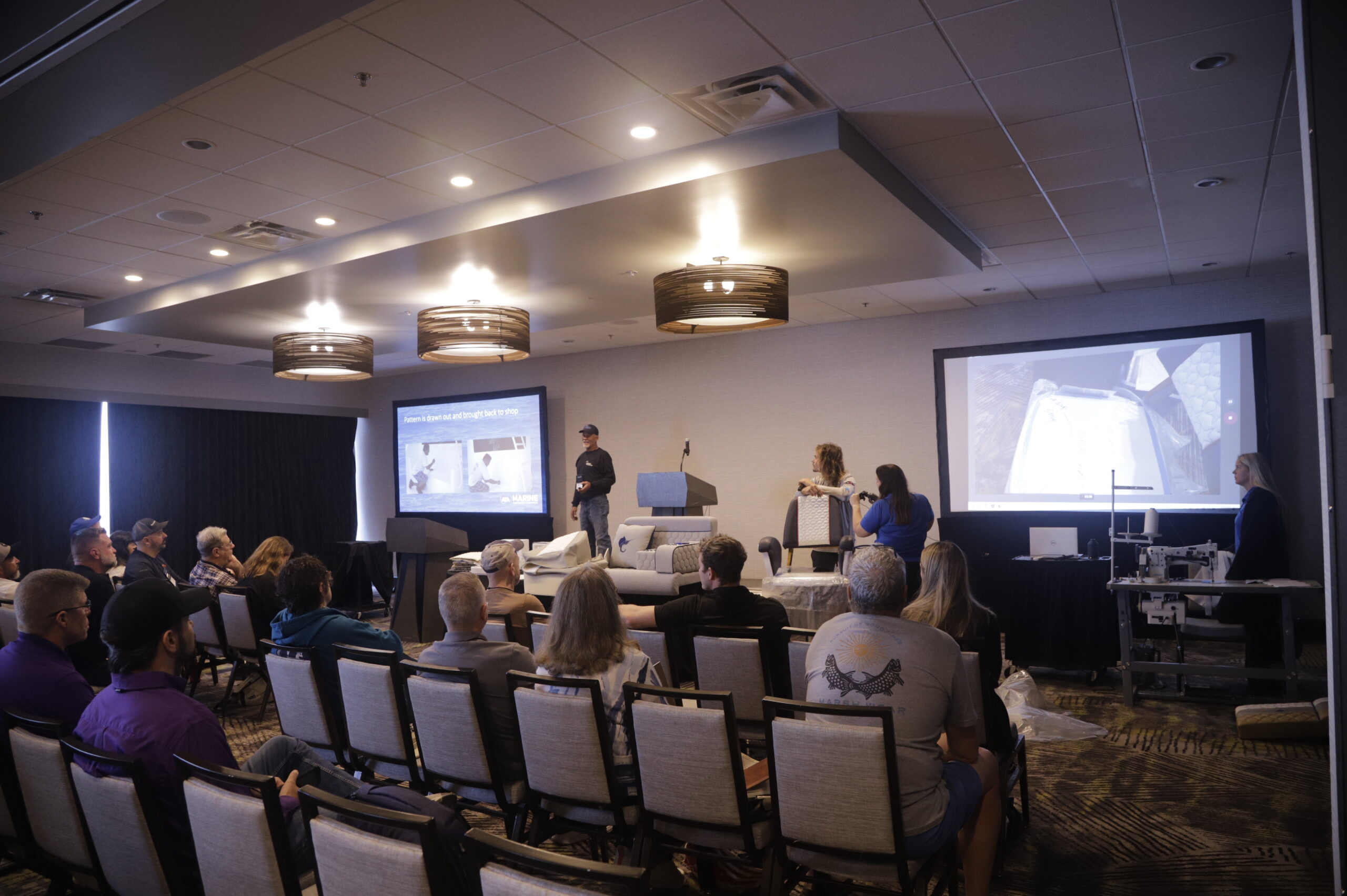 Conference attendees seated in rows watch a speaker present on stage, with a large screen displaying marine fabrication techniques.