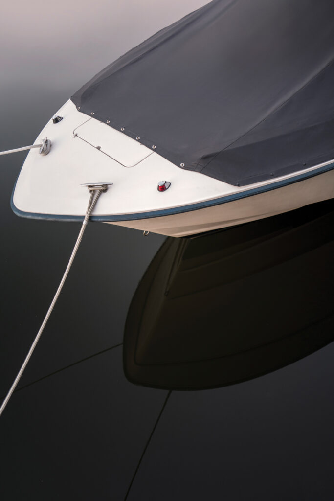 Bow of a boat with a sleek black cover, reflecting in calm waters, surrounded by subtle shades of gray and a hint of light in the background.