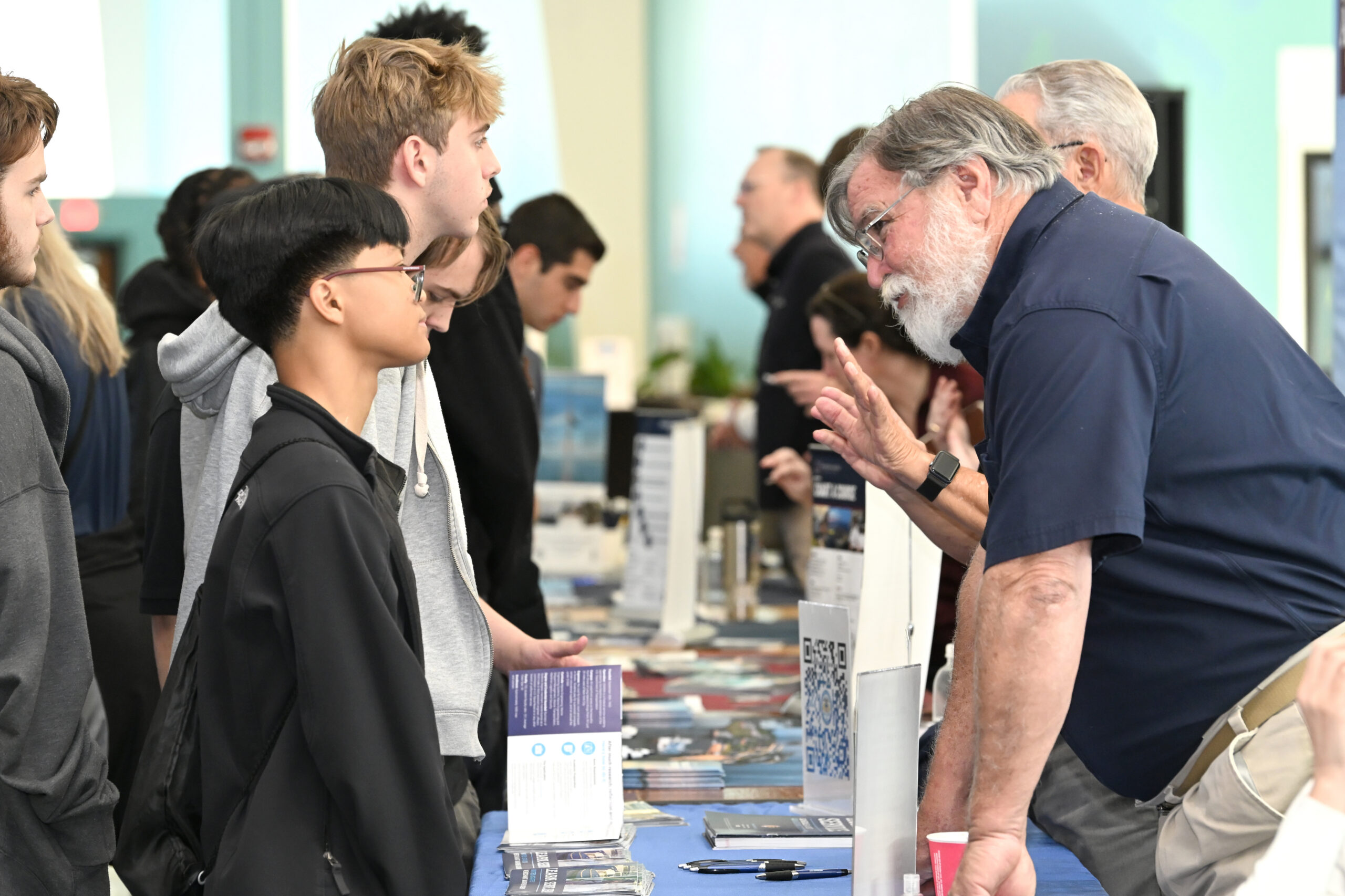 A business owner discusses opportunities with students at a table filled with brochures and resources during a mentorship event.