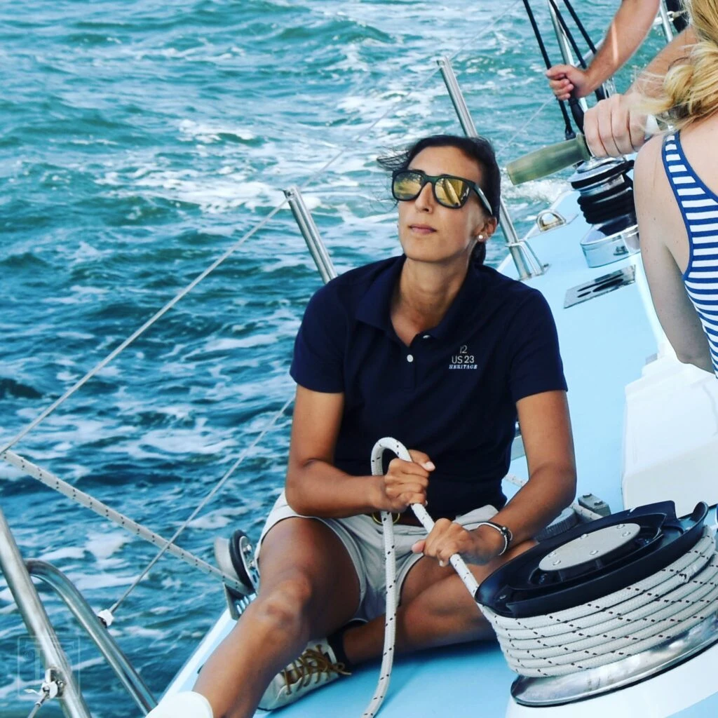 A woman in a polo shirt and sunglasses sits on a sailboat, managing ropes while others help steer on a sunny day.