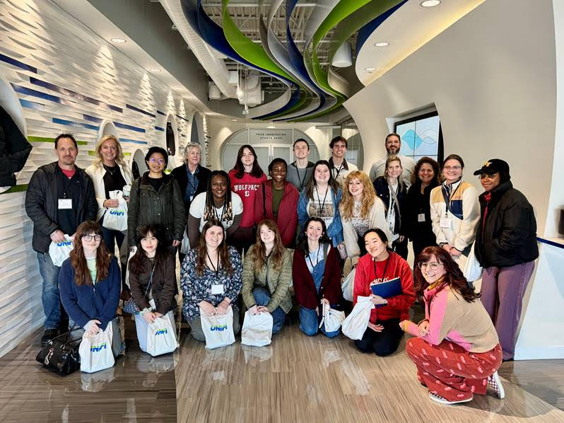 Group of diverse people posing together at UNIFI, each holding bags with the company logo. They took part in the inaugural Spring Break Textiles Trek. Students were from NC State's Wilson College of Textiles and WVU. Amy Bircher Bruyn and MMI Textiles, back row, second from left, was the driving force behind the event.