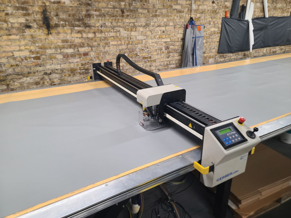 A digital cutting machine with a sliding mechanism on a grey work surface, set against a textured brick wall.