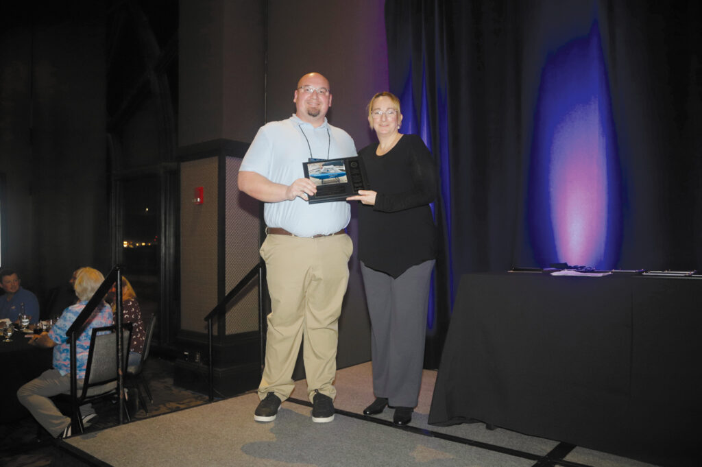 A man and woman stand on a stage, the man holding an award plaque. A black table is nearby, and colorful lighting illuminates the backdrop.