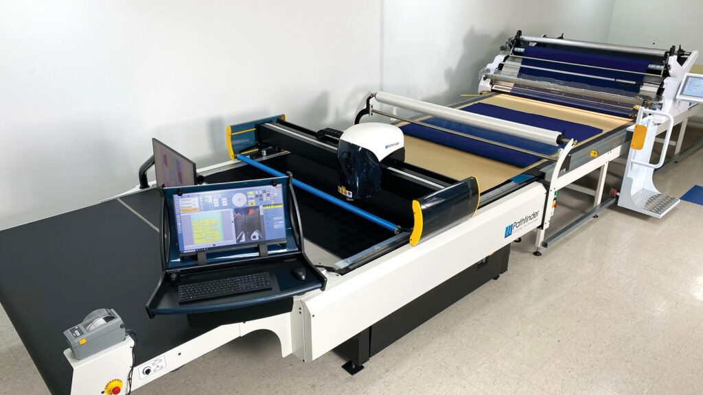 A cutting machine with a computer interface, featuring a black cutting table and a large roll of material on the side, in a bright workspace.