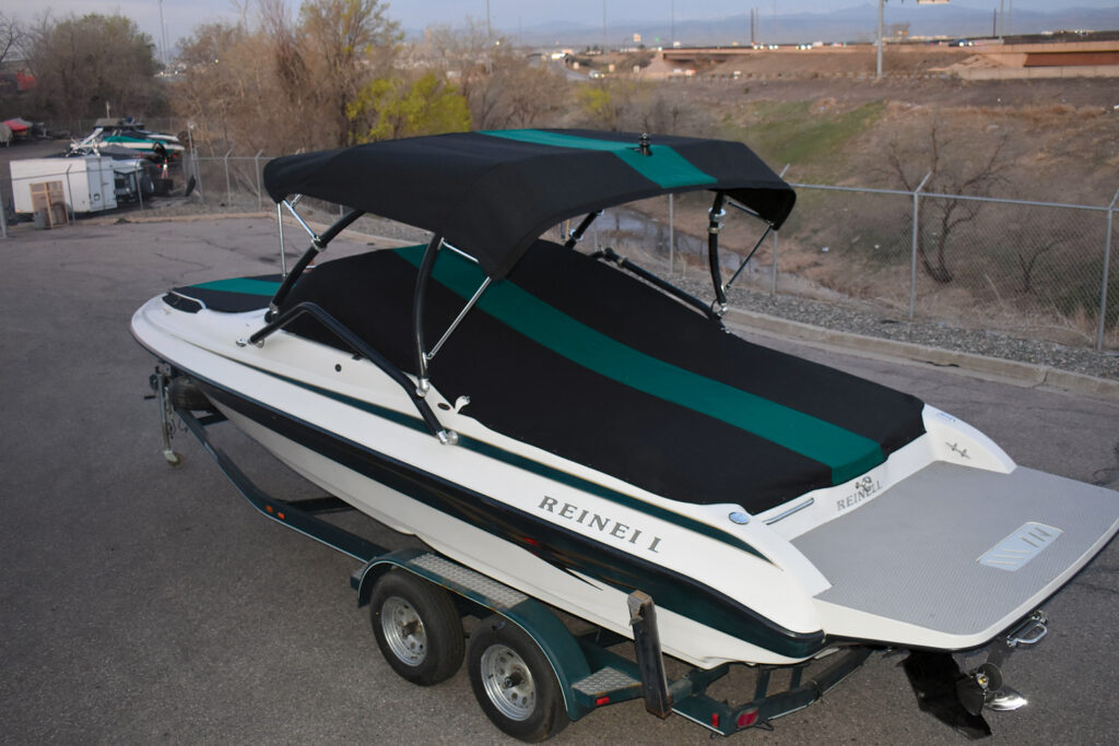 A sleek, black and teal-covered boat with a canopy parked on a trailer, set against a background of bare trees and distant hills.