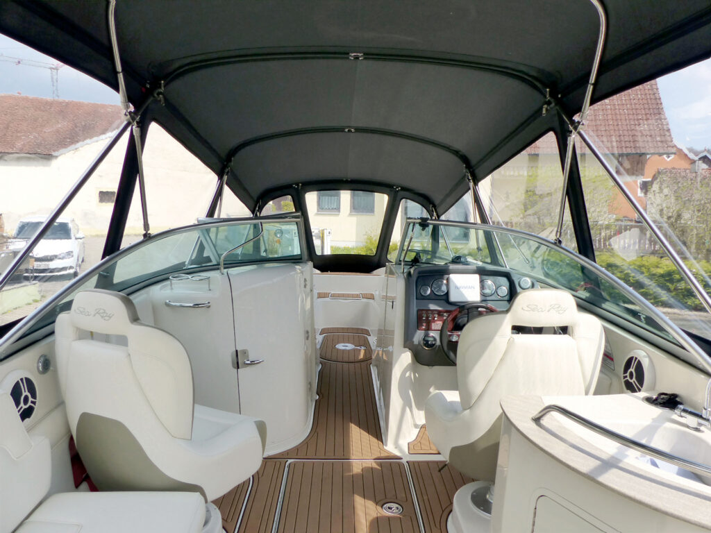 Interior view of a Sea Ray boat featuring white seating, a sleek control panel, and a canopy, with a wooden deck and surrounding buildings.