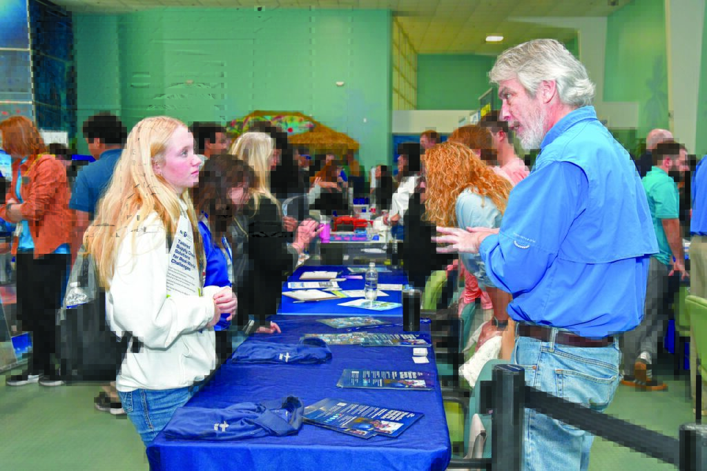 A crowded event scene featuring a conversation between a man in a blue shirt and a woman, with tables displaying brochures and merchandise.