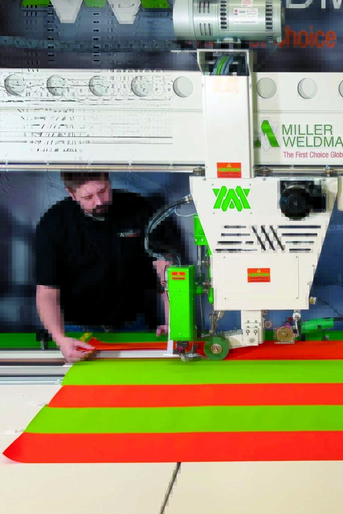 A man operates a large welding machine, creating vibrant orange and green fabric strips, set against a sleek, modern workspace background.