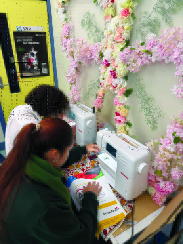 Two individuals are using sewing machines amidst a backdrop of pink and white flowers, focused on fabric projects on a colorful table.