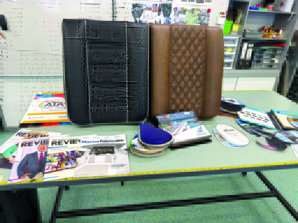 Two fabric seat covers — one black with vertical stitching and the other brown quilted — are displayed on a table with magazines and swatches.