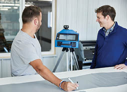 MF_FeaturedImages_254x184px2 Two men discuss a project at a workspace, with one using a marker on a material and a blue equipment setup visible in the background.