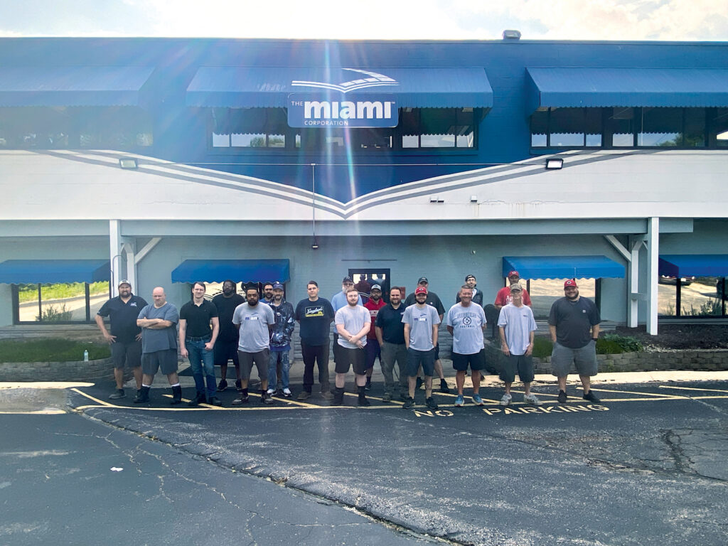 Group of diverse individuals standing in front of a building with blue awnings and a "Miami" sign, lined up in casual clothing.
