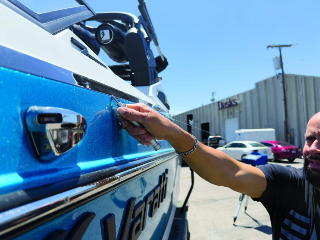 A person adjusts a tool on a blue and silver boat exterior, with a warehouse and parked vehicles in the background.