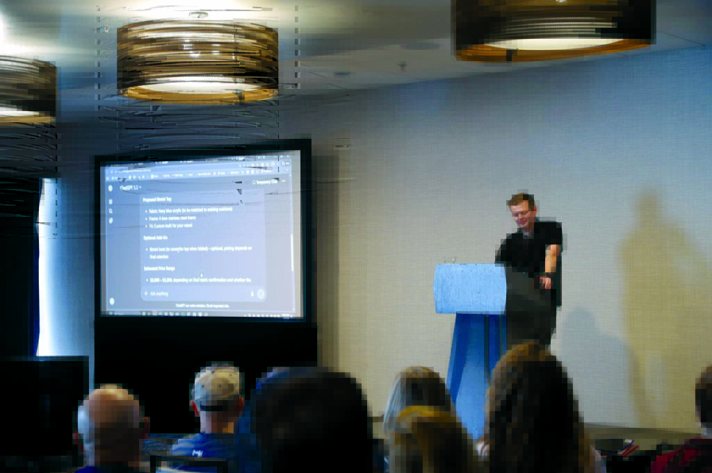 Presenter standing at a podium, speaking to an audience. A large screen displays presentation content, with overhead lighting illuminating the scene.