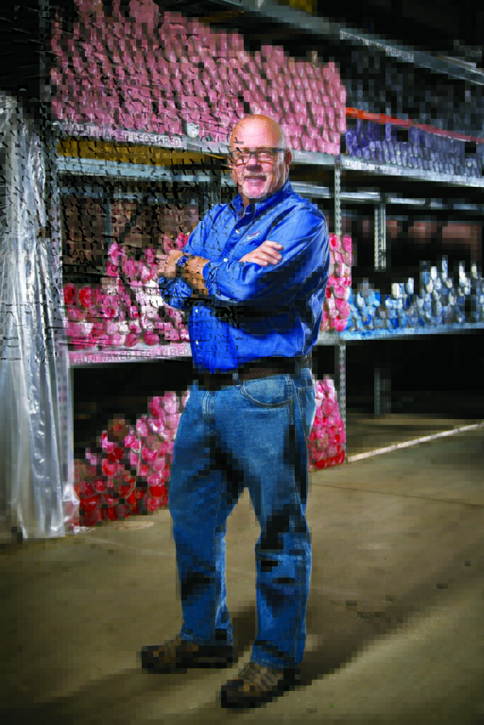 A man in a blue shirt stands confidently with arms crossed, in front of shelves filled with colorful marine fabrication patterns.