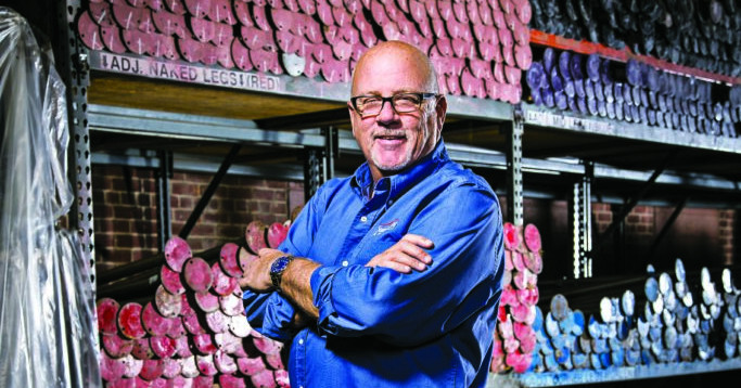 CMB_140813_SteveFrost-1130-EditF-2B-683x1024_WP A smiling man in a blue shirt stands with arms crossed in front of colorful, organized shelves filled with marine fabrication materials.